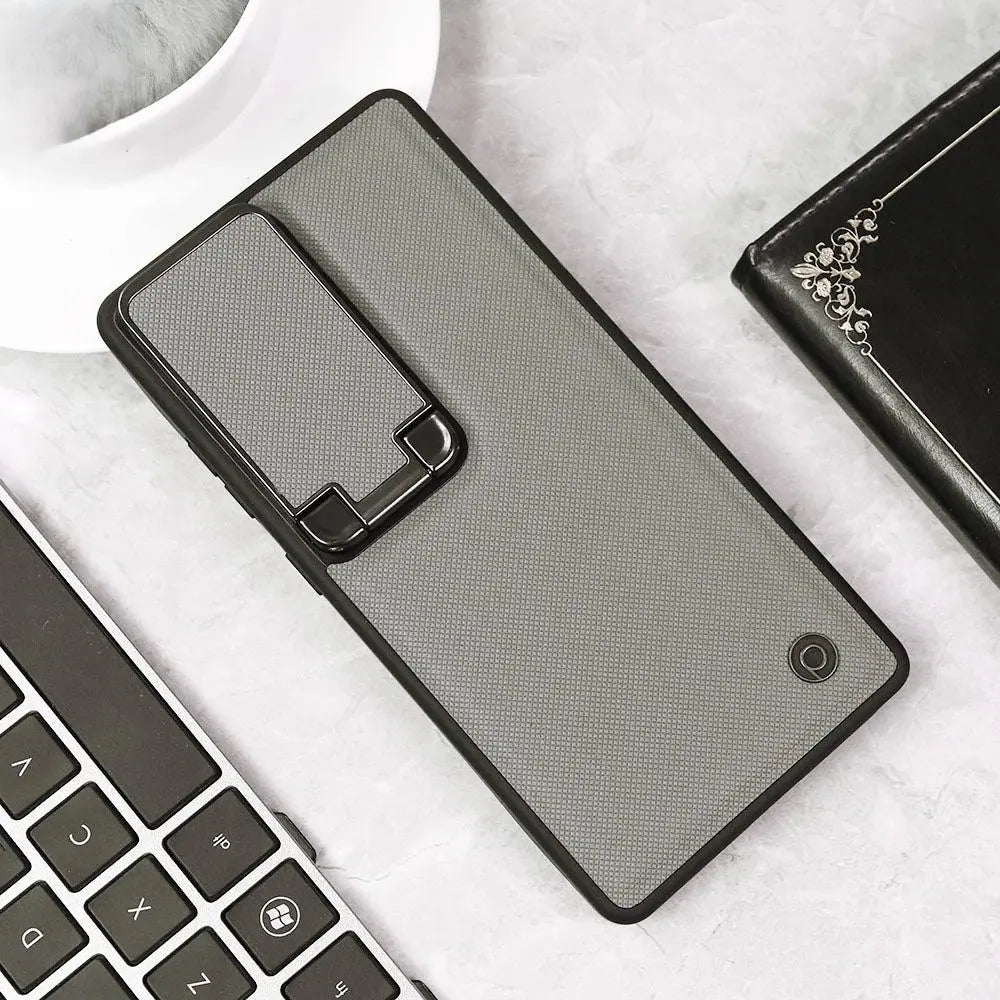 A gray textured phone case with a black kickstand hinge, shown on a light marble surface with a keyboard, coffee cup, and wallet nearby.