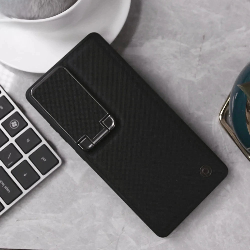A black textured phone case with an integrated kickstand, shown from a top-down angle on a gray marble surface next to a keyboard and a white coffee cup.