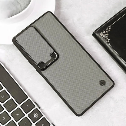 A gray textured phone case with a black kickstand hinge, shown on a light marble surface with a keyboard, coffee cup, and wallet nearby.